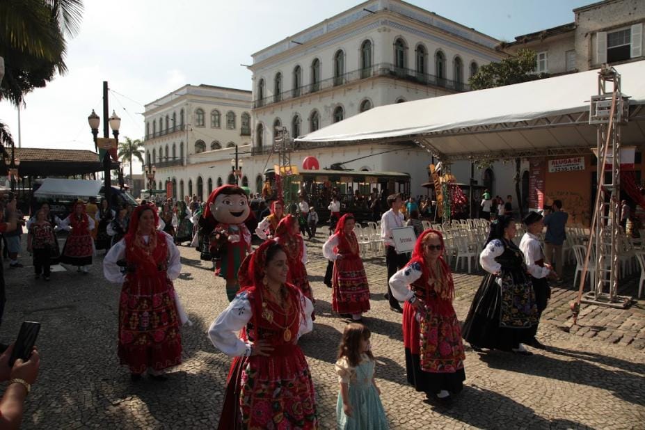 Cortejo de ranchos folclóricos com mulheres em trajes típicos vermelhos e mascote gigantone caminham pelo Centro Histórico de Santos na Festa de Portugal.