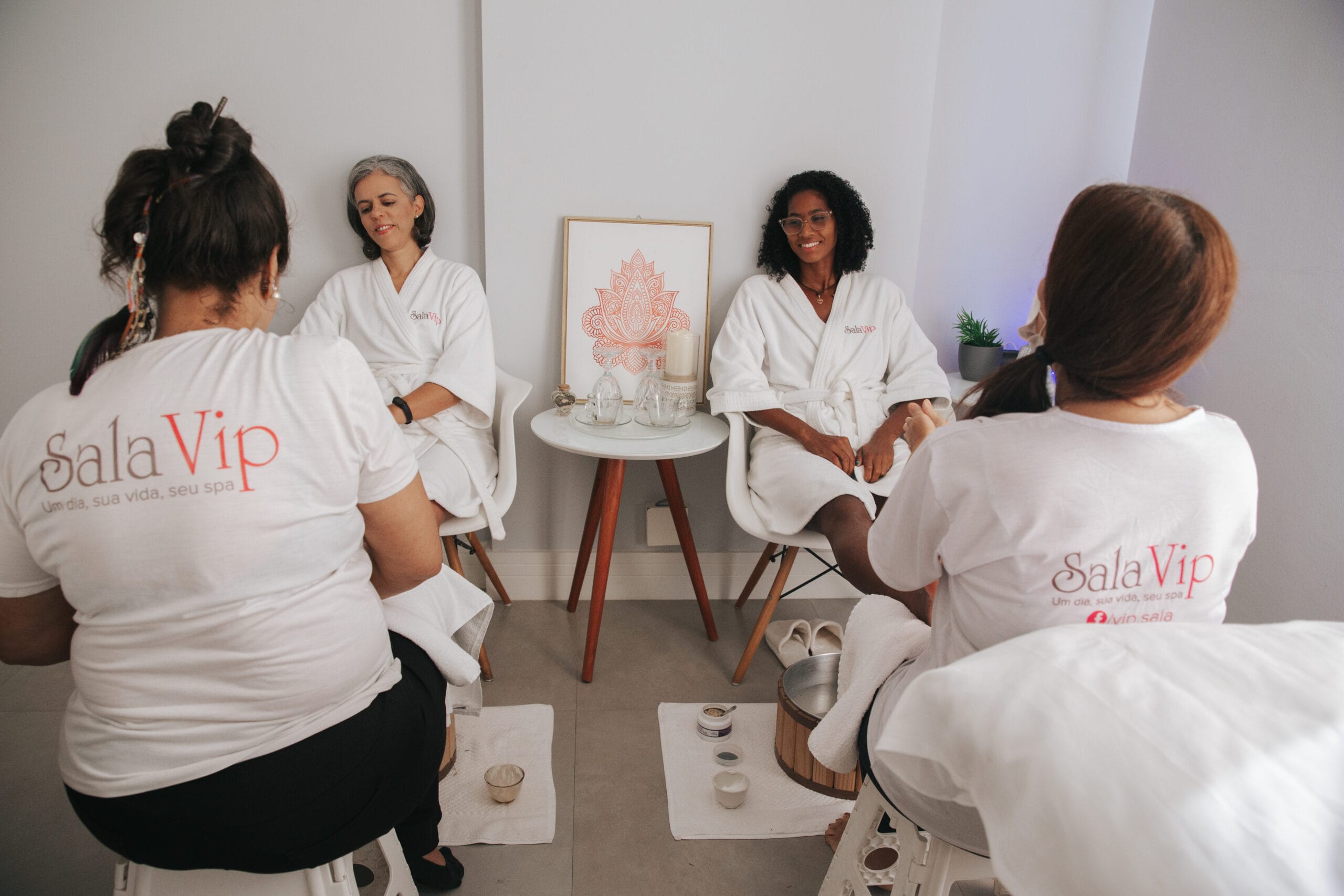 Duas clientes de roupão branco recebendo escalda-pés de profissionais da Sala Vip Spa durante um Day Spa em grupo.