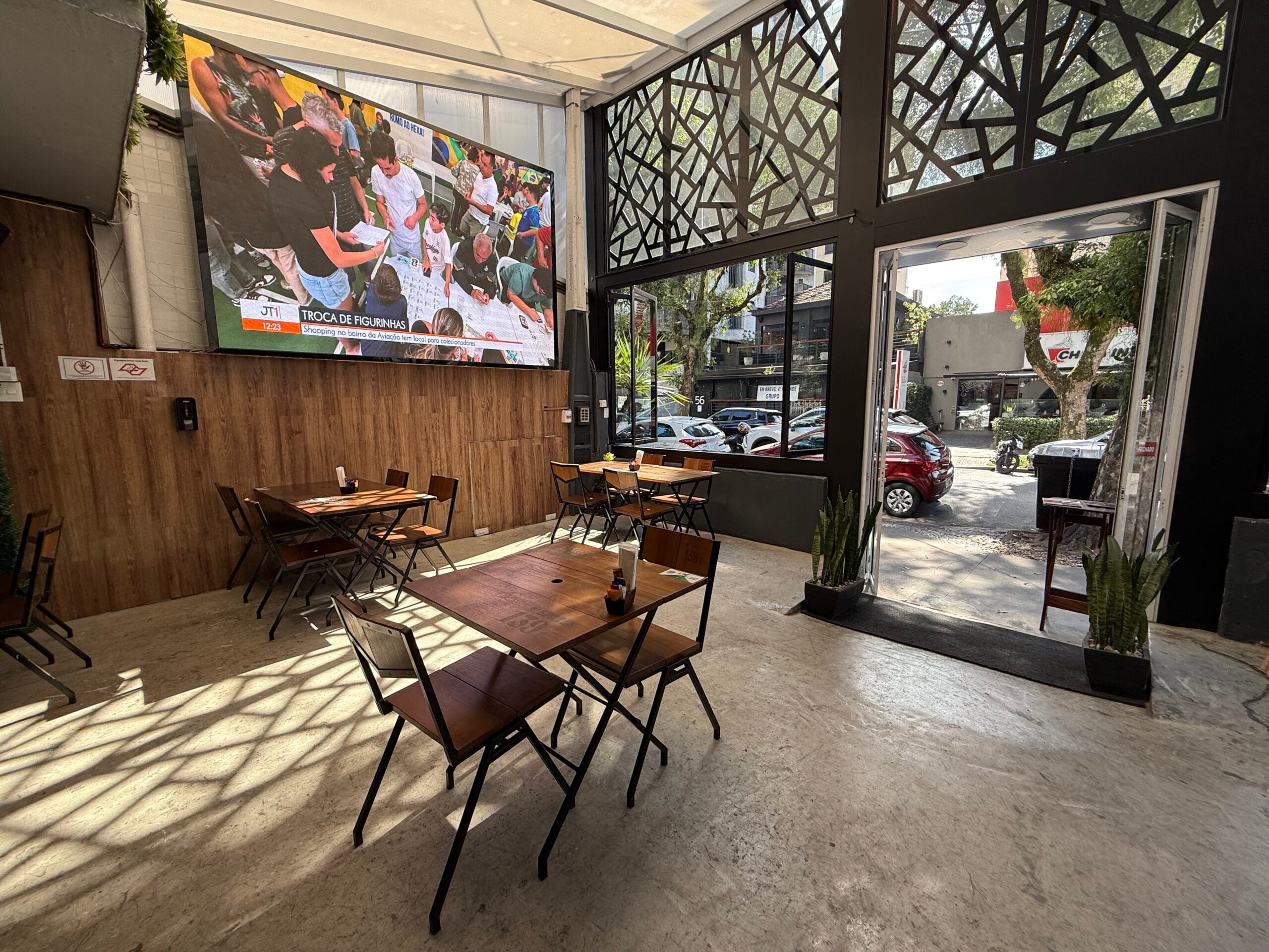 Salão frontal do Zora Bar com mesas, telão de TV e luz natural pela porta principal