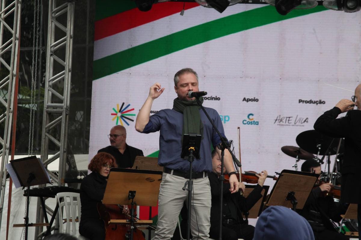 Cantor se apresenta com orquestra no palco da Festa Italiana de Santos, com o painel do evento ao fundo